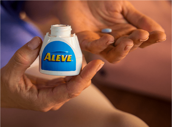 A hand holds out an Aleve® pill and another holds the Aleve® bottle
